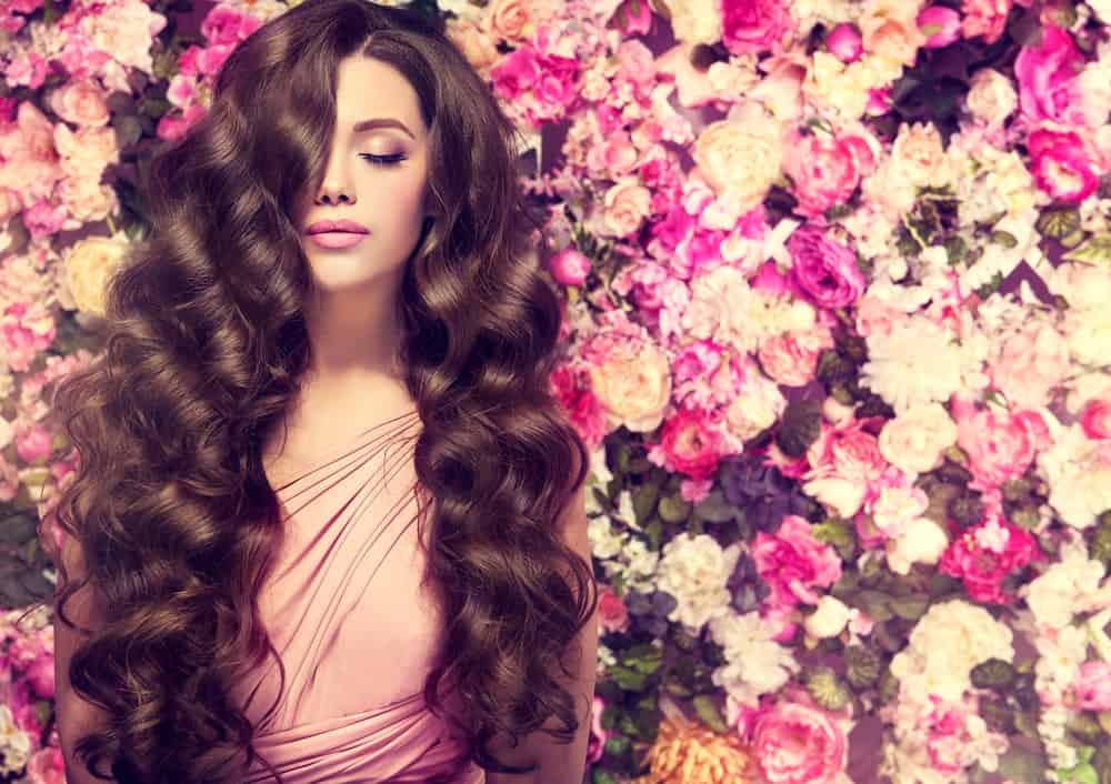 Brunette girl with long and shiny wavy hair on a background wall of flowers.