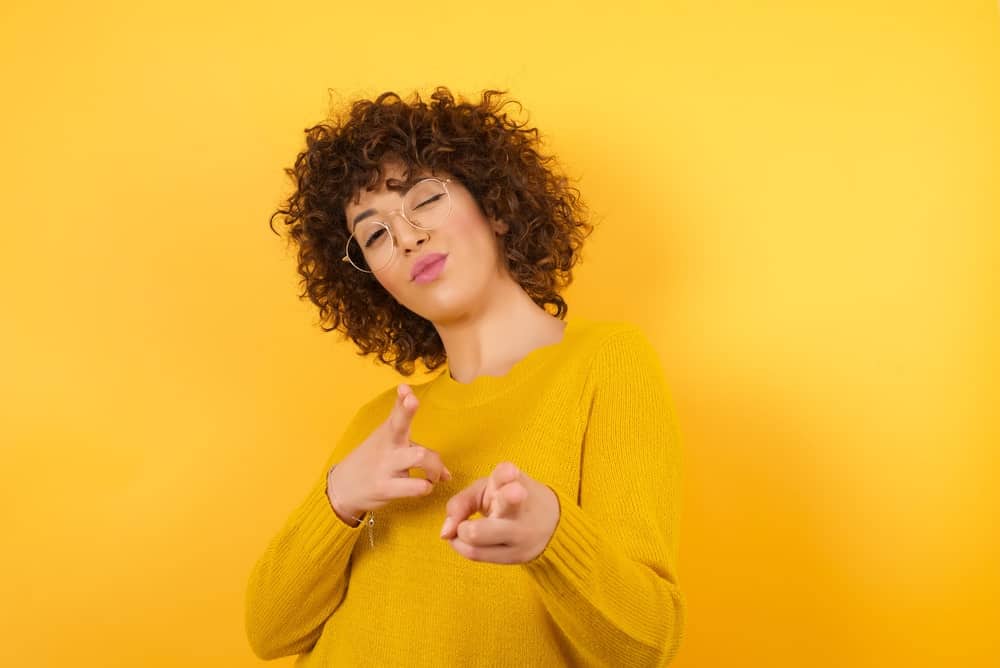 Woman with short curly hair and eyeglasses points with finger pistols to the camera against a yellow background.