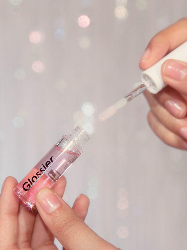 Close-up shot of hand (wearing clear nail polish) about to dip the wand into Glossier Lip Gloss, against a light-colored blurred sparkly background