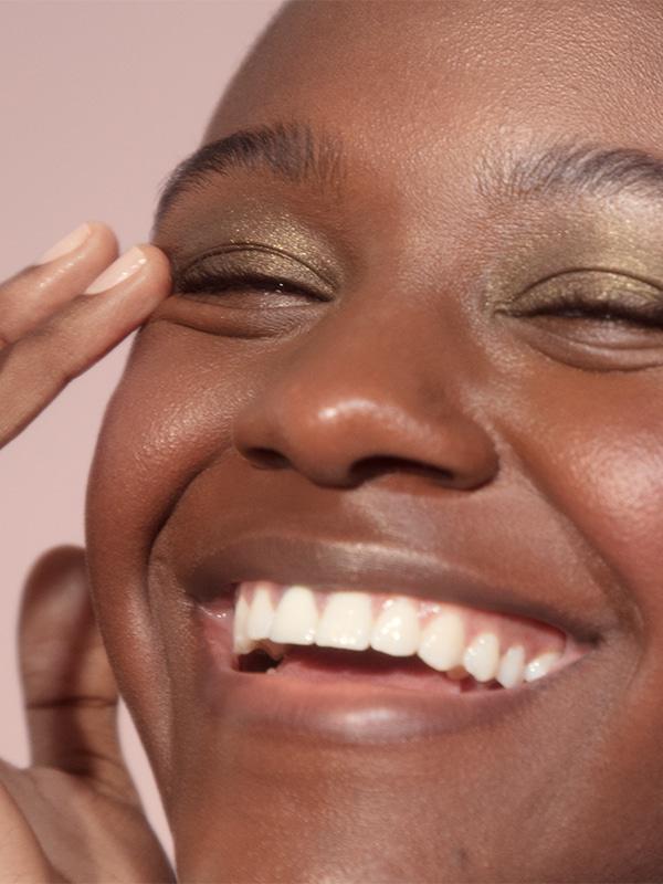 Close-up shot of woman smiling against pale pink background and wearing Glossier Lidstar in 